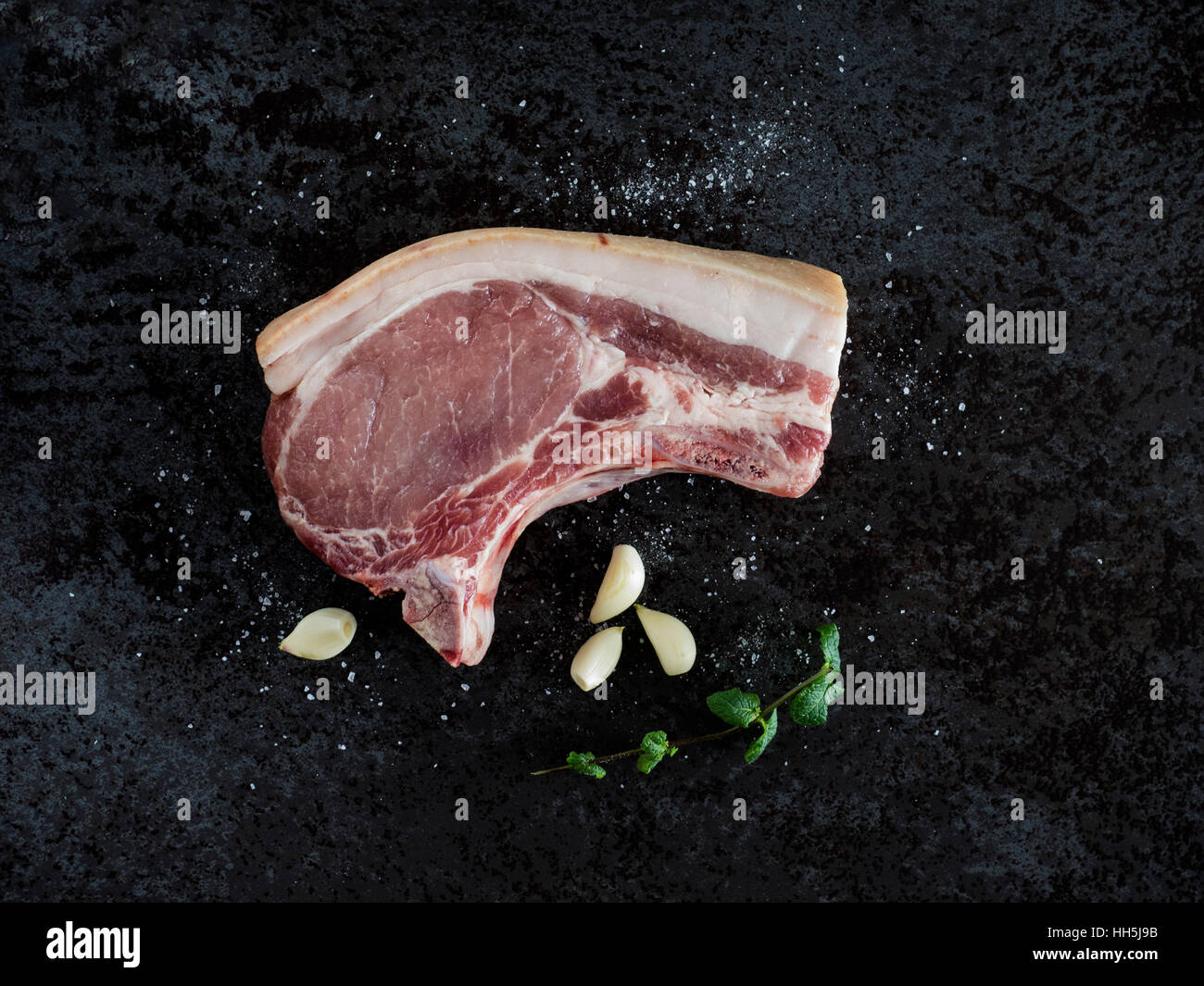 Rohes Schweinefleisch Chop, Knoblauch, Minze und Salz auf schwarzem Stein Stockfoto