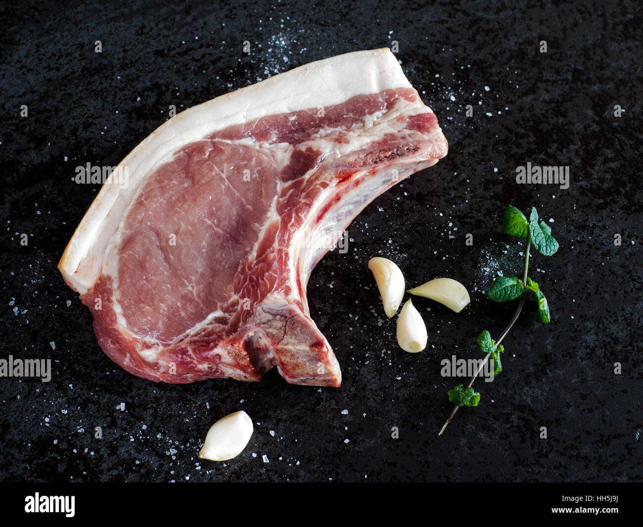 Rohes Schweinefleisch Chop, Knoblauch, Minze und Salz auf schwarzem Stein Stockfoto