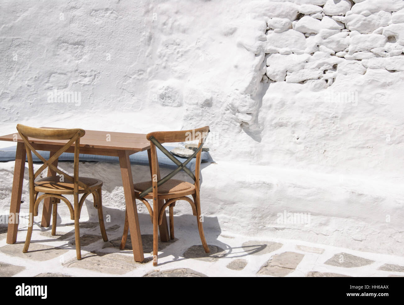 Ein leerer Außentisch mit zwei Stühlen steht in der griechischen Sonne auf einer weiß getünchten Terrasse. Stockfoto