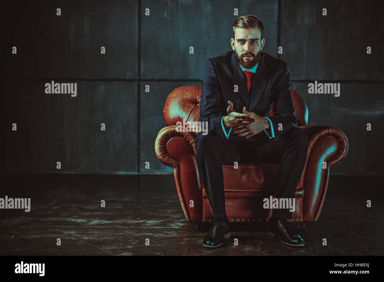 Junge hübsche Geschäftsmann mit Bart im schwarzen Anzug auf Stuhl sitzend und im Gespräch mit Lautsprecher. Film-Stil-Retro-Farben-Effekt. Stockfoto