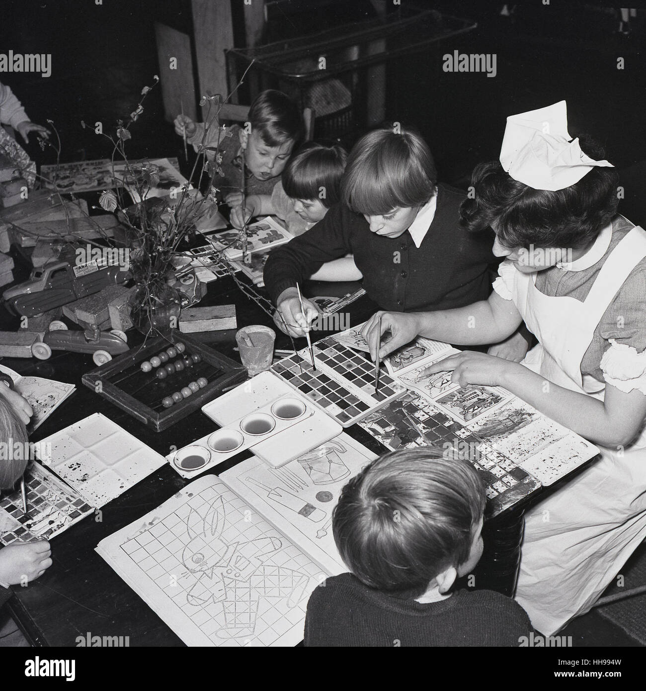 1950, historische, eine uniformierte weibliche Krankenschwester in ein Kinderkrankenhaus sitzen, helfen die kleinen Kinder mit ihren Aquarellmalerei. Stockfoto