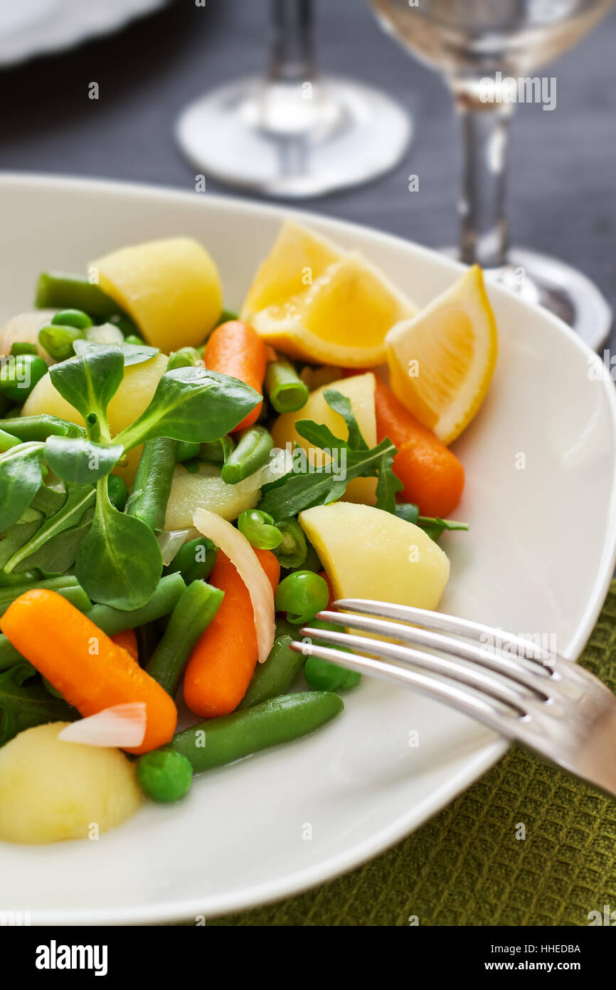 Salat mit Baby-Karotten, Erbsen, Zwiebeln und Kartoffeln serviert auf weißen Teller auf schwarzem Hintergrund aus Holz Stockfoto