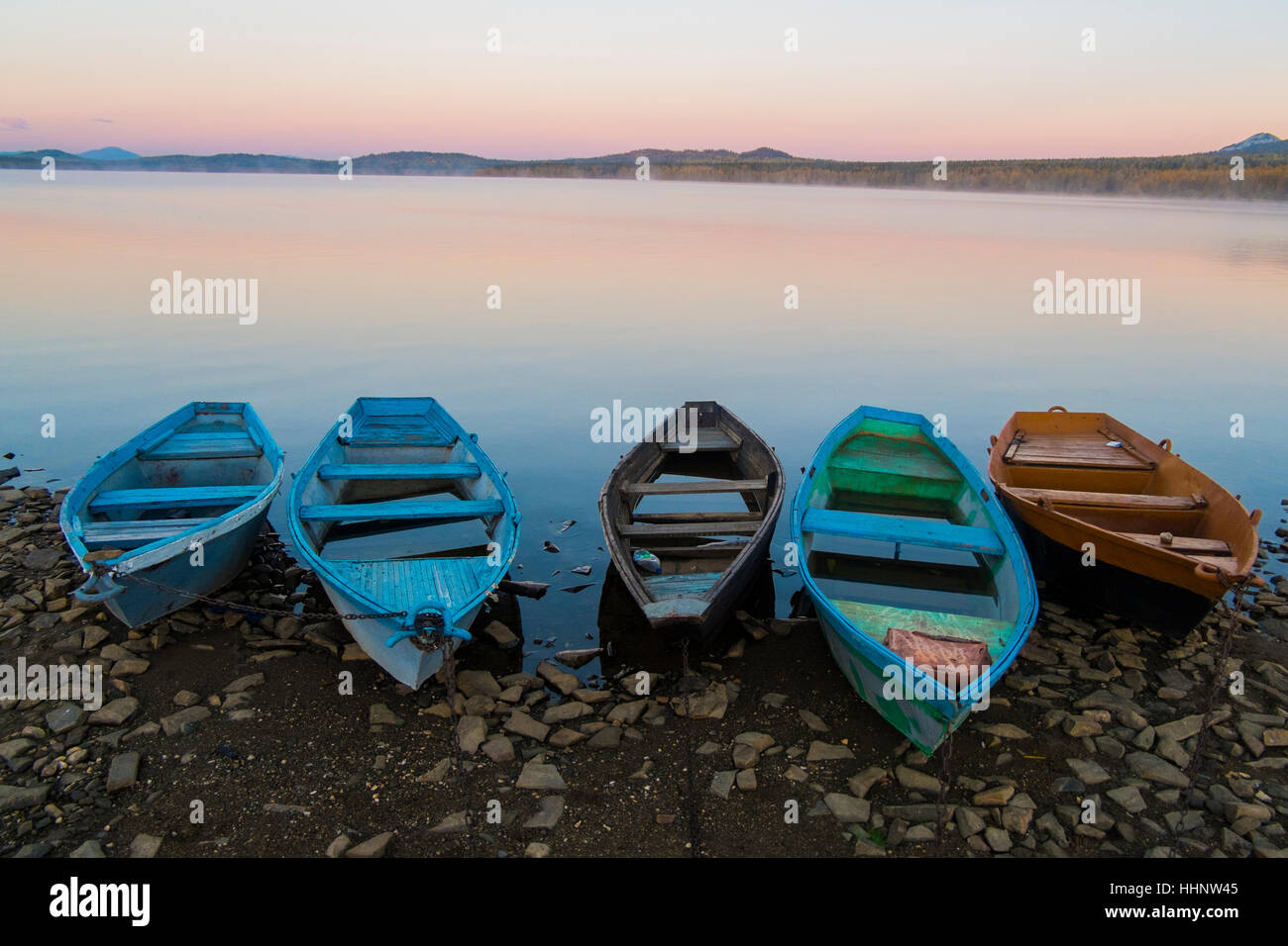 Fünf leere Ruderboote am felsigen Ufer des Sees bei Sonnenuntergang Stockfoto