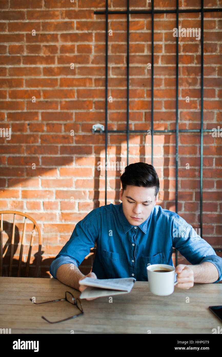 Junger Mann, Zeitung lesen, Kaffee zu geniessen, an einem Holztisch im Hipster-Café sitzen und blickte, Telefon und Gläser auf Tisch Stockfoto