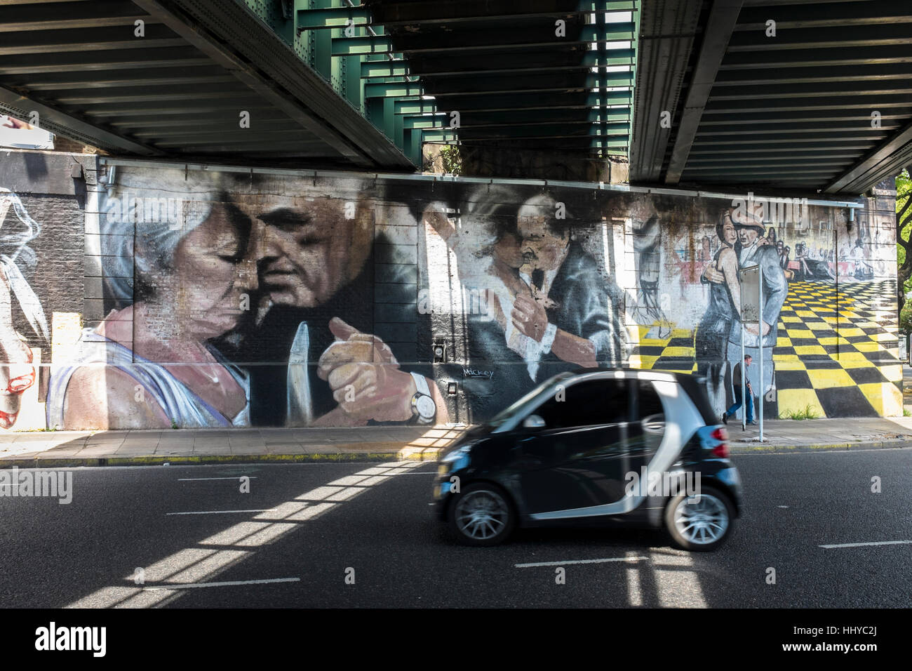 Ein Auto fährt unter einer Eisenbahnbrücke wo gestrichene Wände zeigen drei Paare tanzen Tango in der Stadt Buenos Aires, Argentinien. Stockfoto