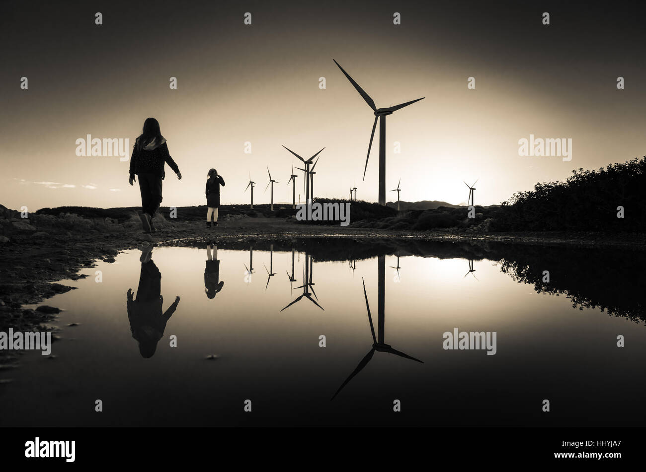 Windkraftanlagen im Windpark mit den Silhouetten von zwei Mädchen, die auf der Wasseroberfläche spiegelt sich bei Sonnenuntergang, Kreta, Griechenland Stockfoto