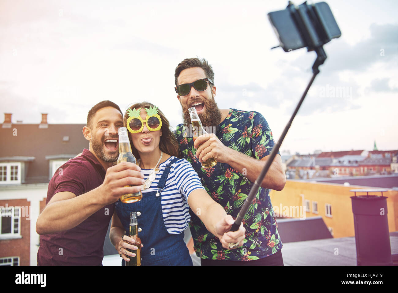 Gruppe von drei Erwachsenen trinken und nehmen Bilder von sich selbst auf Dach mit Selfie Stick auf Dach Stockfoto
