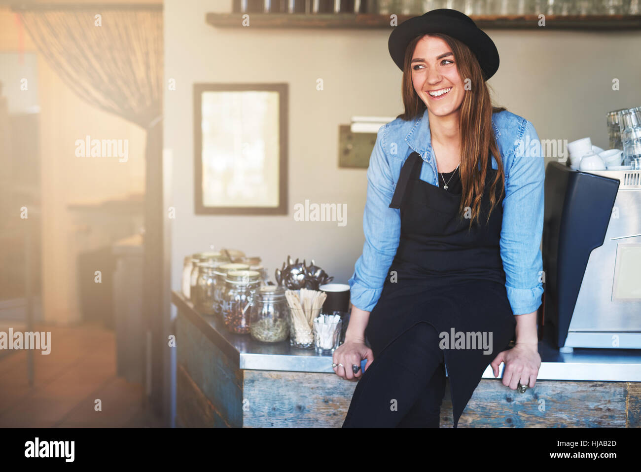 Junge Frau in schwarzer Schürze an Kaffee Schreibtisch saß und lächelnd Weg auf Grund des Café im Haus. Stockfoto