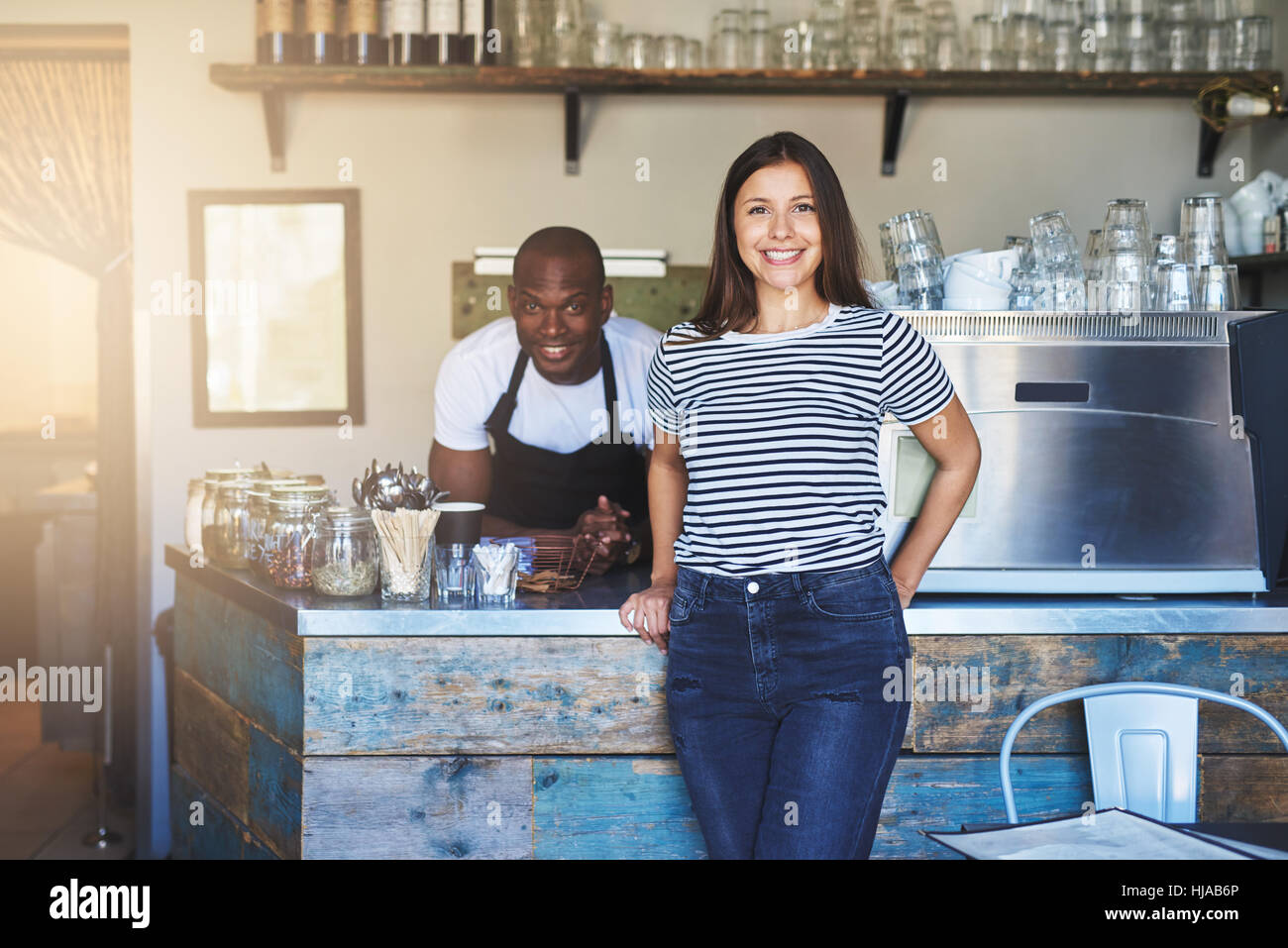 Mädchen lächelnd an Kamera stehend in der Nähe von Kaffee zu machen, im Café mit schwarzen männlichen Arbeiter hinter stehen. Stockfoto