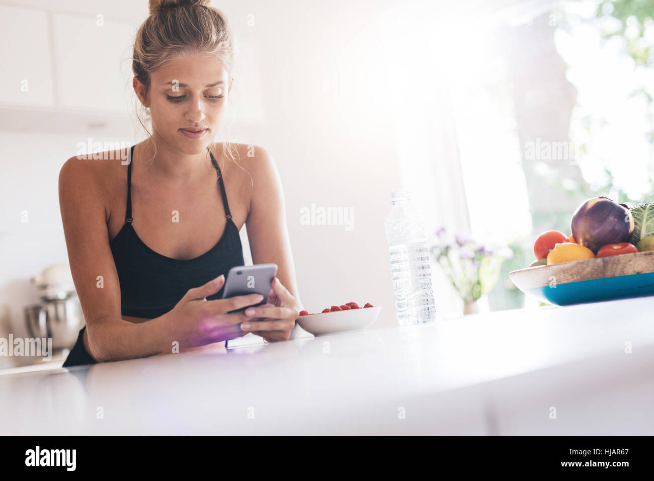 Junge Frau mit Handy in der Küche beim Frühstück Essen. Schöne weibliche lesen SMS-Nachricht auf ihr Smartphone. Stockfoto