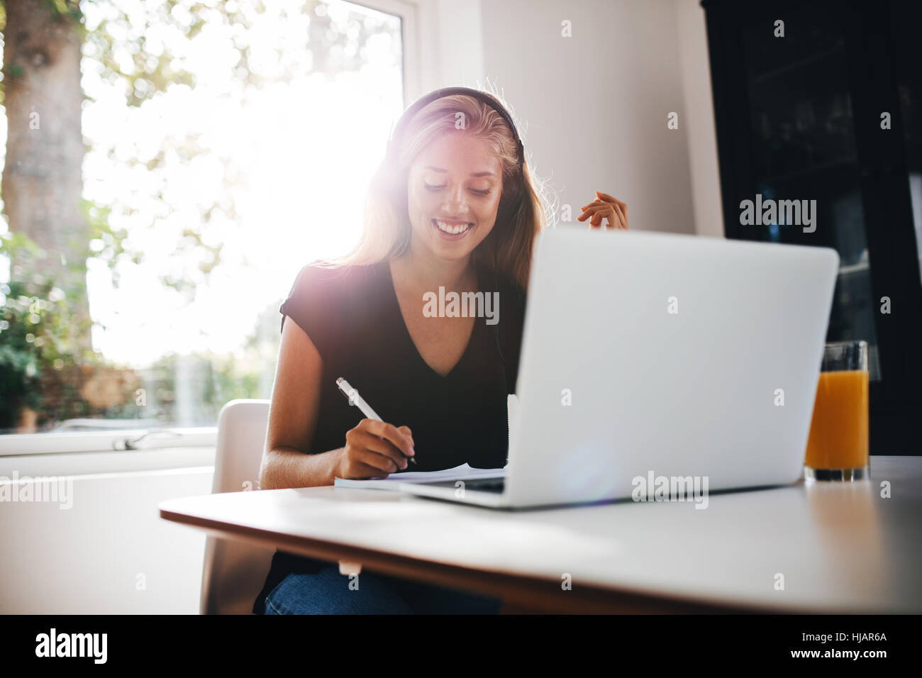 Innenaufnahme der glückliche junge Frau studieren in Küche. Weibchen mit Kopfhörer mit Laptop am Tisch auf Buch schreiben. Stockfoto