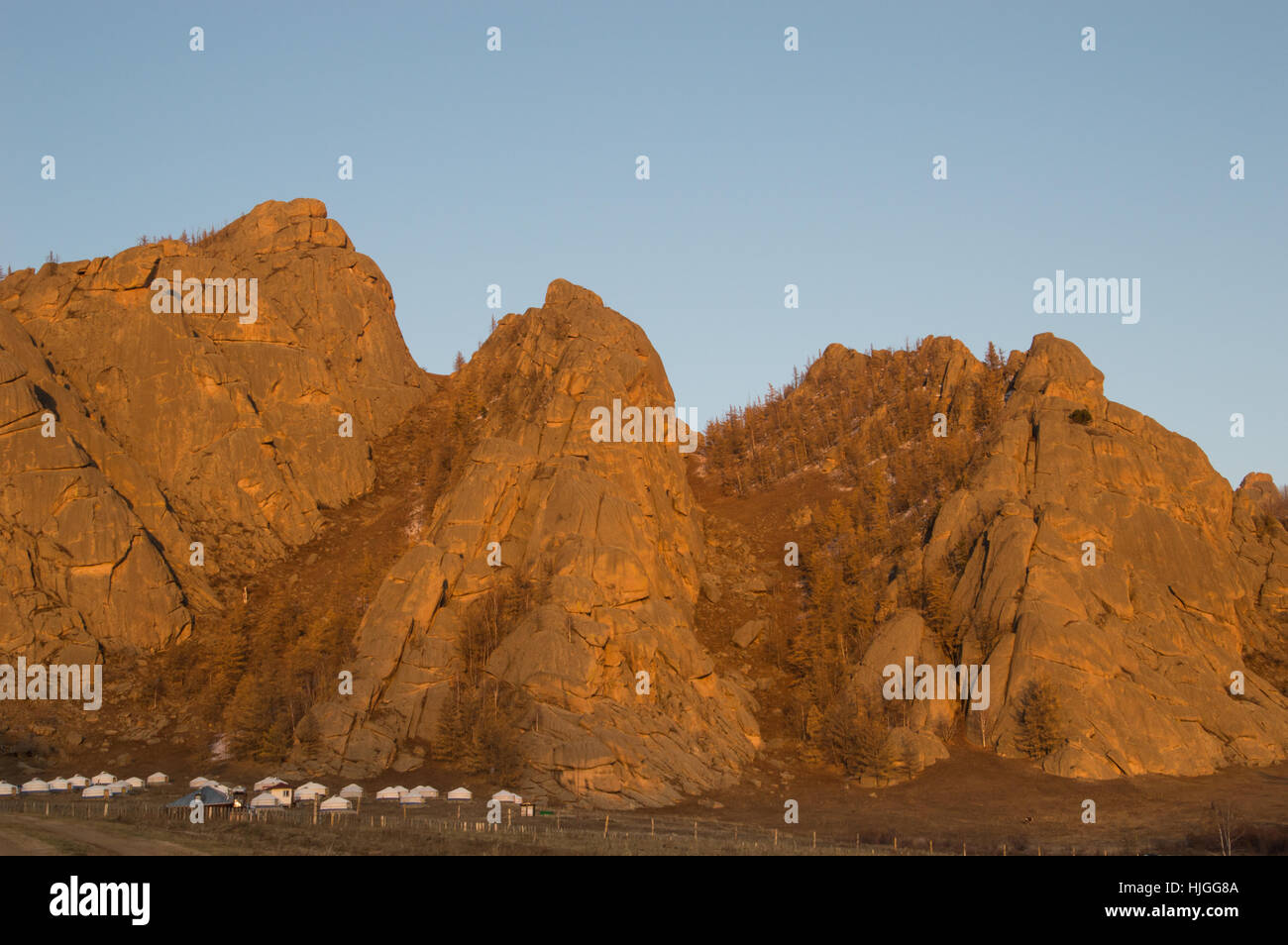 Ein Lager mit mehreren weißen Jurten am Fuße der schroffen Stein Bergen, die in der untergehenden Sonne golden sind. Das Hotel liegt in der Mongolei. Stockfoto