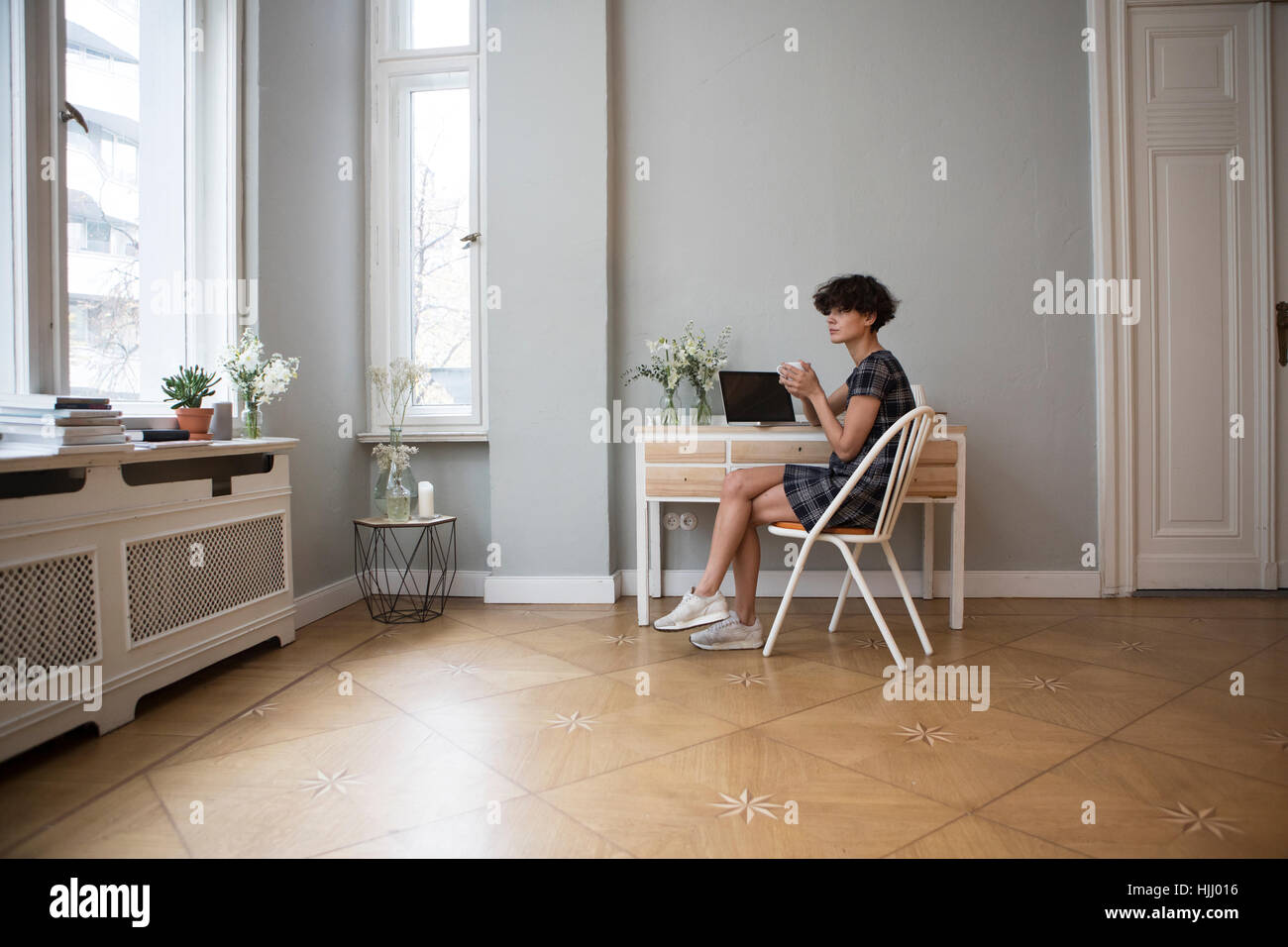 Junge Frau sitzt am Schreibtisch zu Hause mit Tasse Kaffee mit Blick durch Fenster Stockfoto