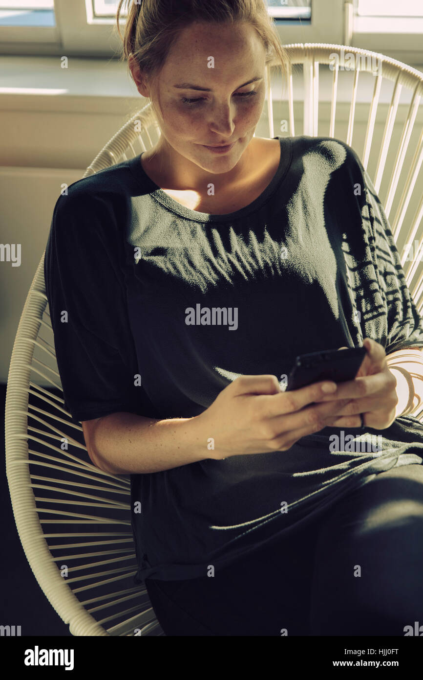 Junge Frau sitzt auf einem Stuhl, Blick auf ihr Handy Stockfoto