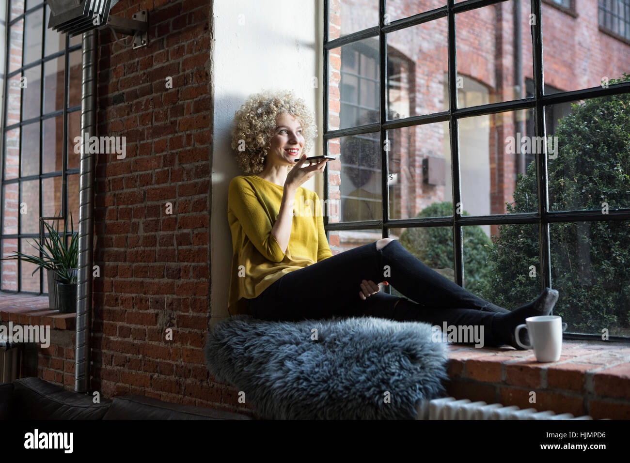 Junge Frau sitzt auf der Fensterbank, in Smartphone sprechen Stockfoto
