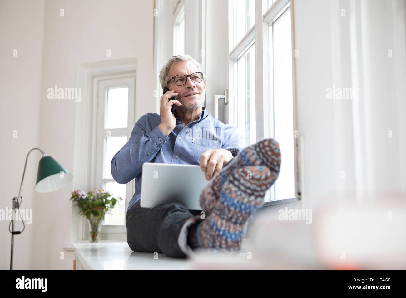 Reifer Mann zu Hause sitzen am Fenster mit Tablet und Handy Stockfoto
