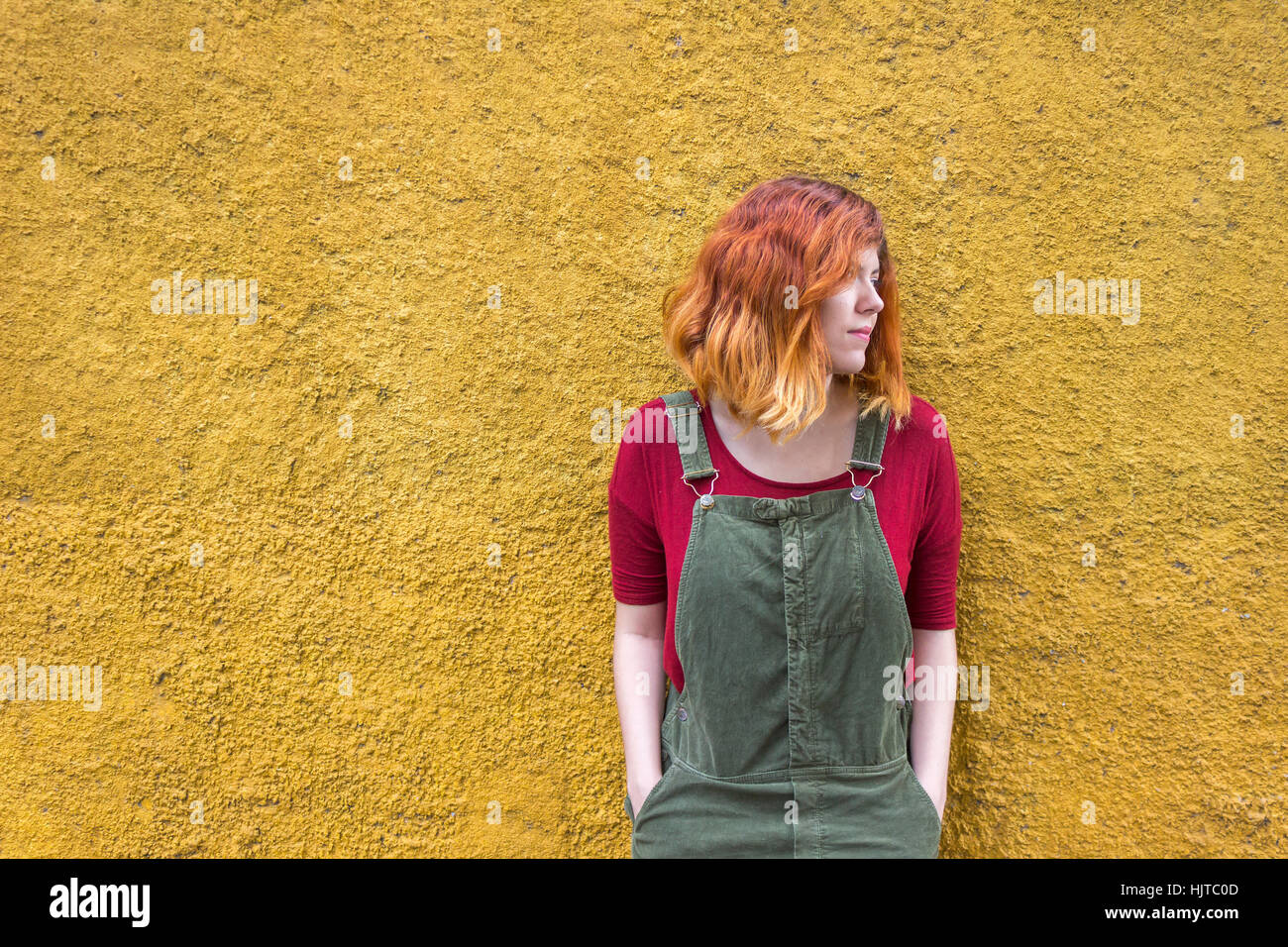 Junge Frau mit roten Kurzhaar und tragen grüne Overalls mit einem roten Hemd Lächeln stehend gegen gelb Stockfoto