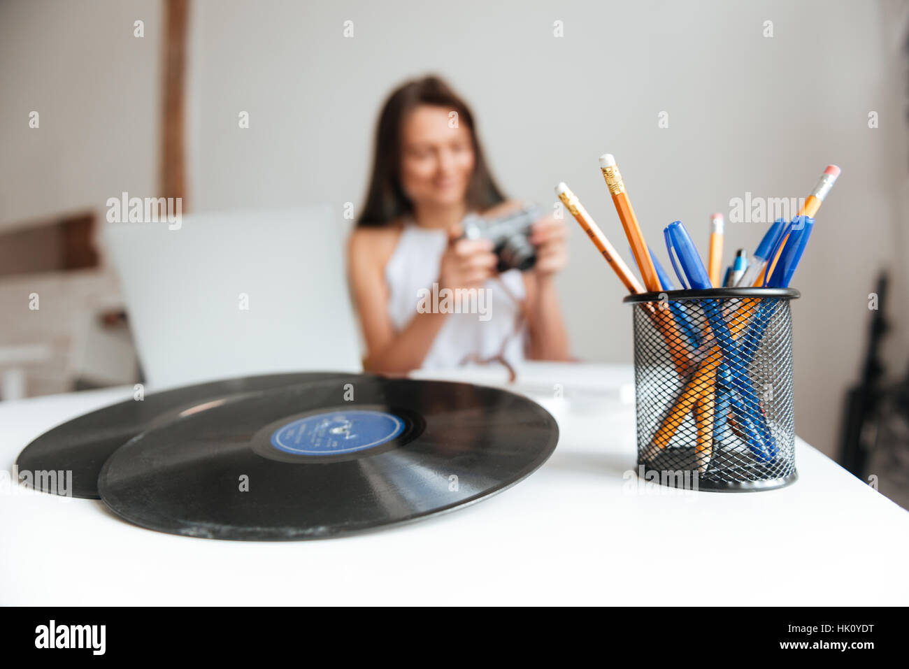 Foto von casual Frau, die auf einem Laptop sitzen auf dem Stuhl im Haus während Kamera arbeitet. Tisch mit Vinyl-Schallplatte und Bleistifte im Mittelpunkt. Stockfoto