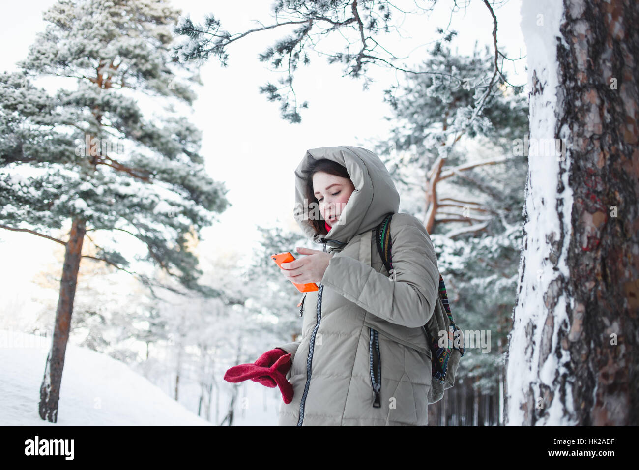 Stets gut gelaunte Frau SMS auf orange Smartphone während einer Reise in den Wald in Brünette Modell tragen warme Winterjacke mit Kapuze, rote Handschuhe Stockfoto
