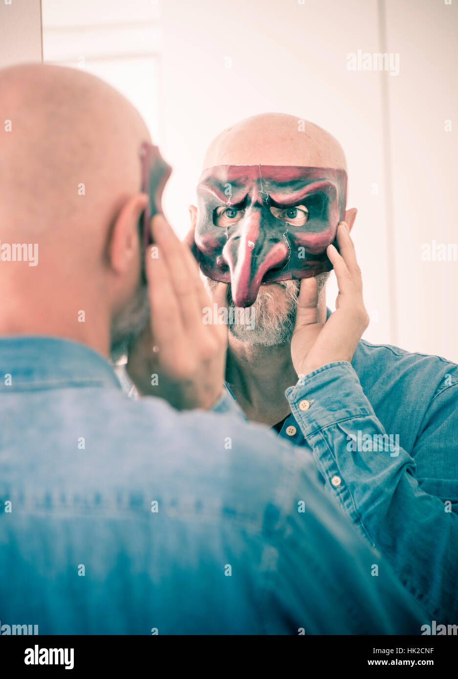 Alte Mann Abdeckung Gesicht mit Karnevalsmaske. Selbst betrachten im Spiegel. Konzept der Überalterung, Selbstbild als eine ältere Person und Identität verbergen. Stockfoto
