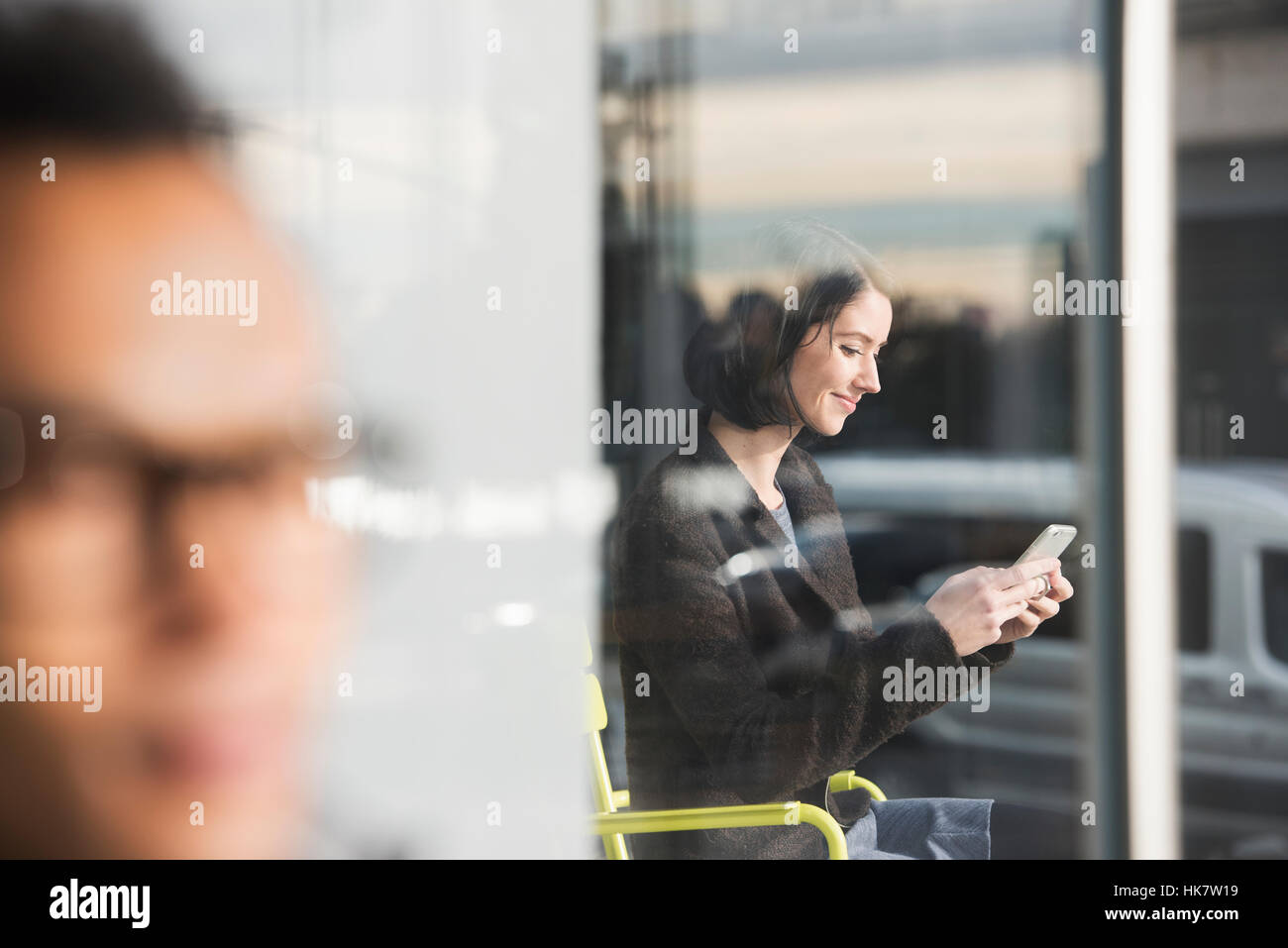 Eine sitzende Frau durch ein Fenster mit Blick auf ein Handy mit einem Out-of-Focus-Mann im Vordergrund zu sehen. Stockfoto