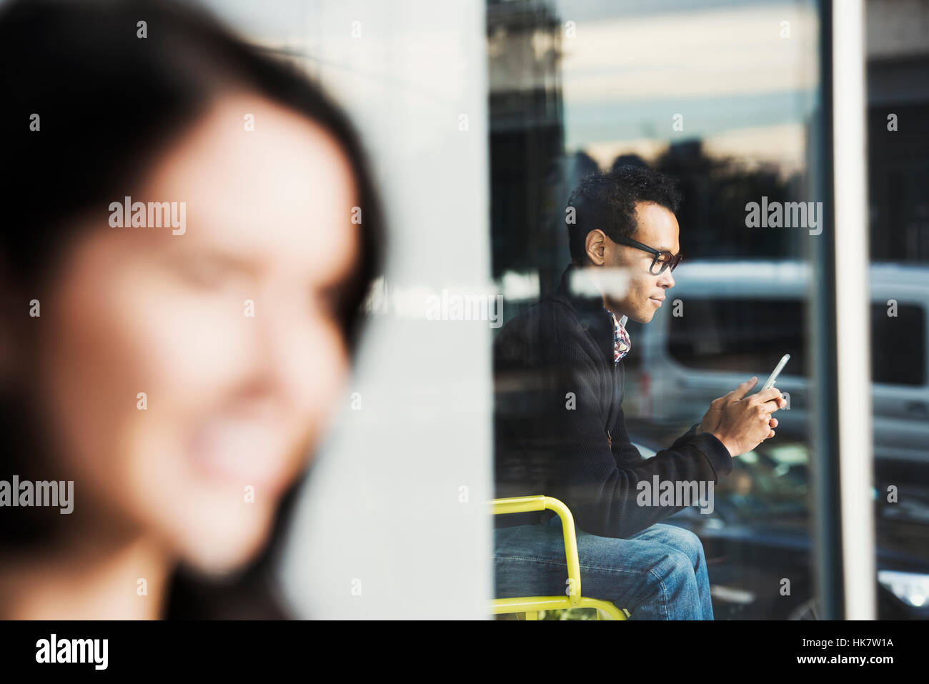 Ein sitzender Mann, gesehen durch ein Fenster mit Blick auf ein Handy mit einer Out-of-Focus-Frau im Vordergrund. Stockfoto