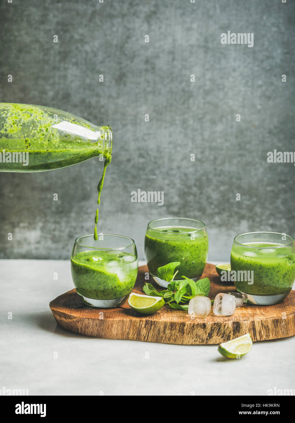 Frischen grünen Smoothie in Gläser und Gießen aus Flasche mit Eiswürfeln, Minze, Limette auf Holzbrett, graue Betonwand im Hintergrund, selektiven Fokus, Stockfoto