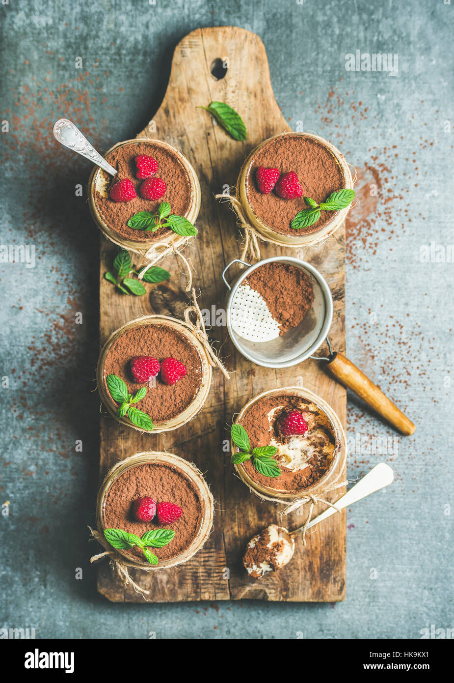 Hausgemachte italienisches Dessert Tiramisu in einzelnen Gläsern mit Minze Blätter, frische reife Himbeeren und Kakaopulver auf rustikalem Holzbrett über graue con Stockfoto