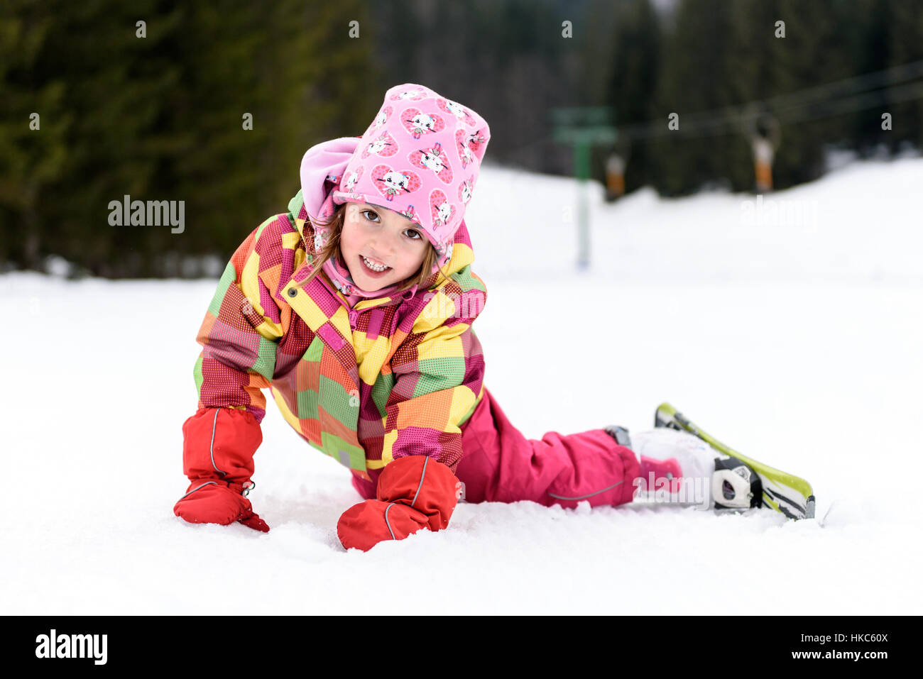 Kleines Mädchen im Winter-Outfit fiel beim Skifahren. Kind ist mit Skiern lächelnd im Schnee liegen. Happy Ski erleben Sie im Resort. Ski-Unfall. Stockfoto