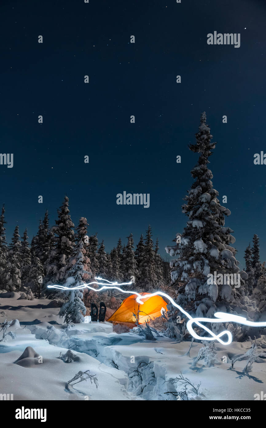 Eine Spur des Lichts führt vorbei an einem Orange Zelt in der Mitte eines verschneiten Evergreen Forest, Schneeschuhe vor dem Zelt, Moonlight Casting Shadows auf Einem ... Stockfoto