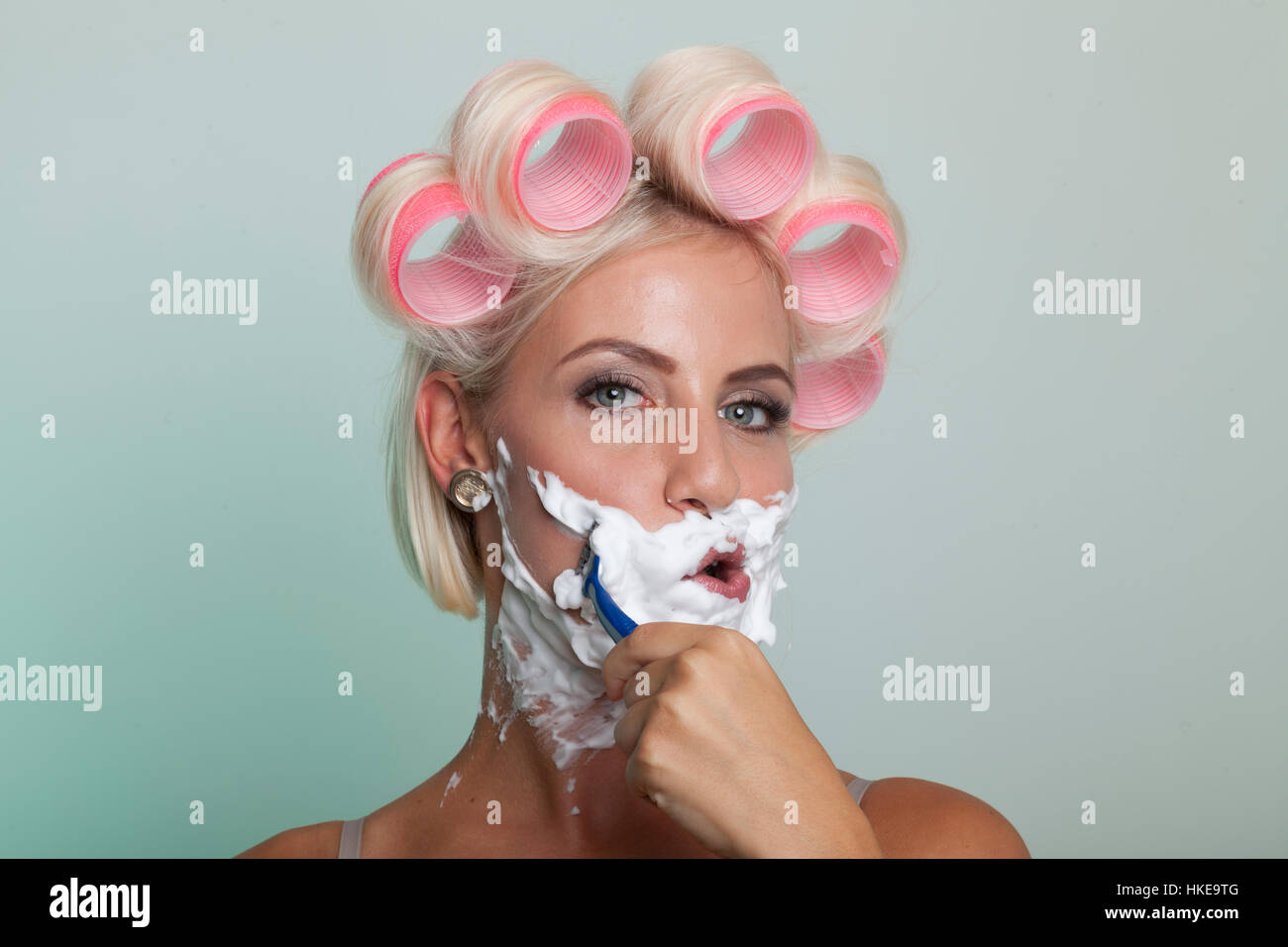 Blonde Frau mit rosa Lockenstäbe auf ihren Kopf und Rasierschaum im Gesicht rasiert ihre Stoppeln Bart Stockfoto