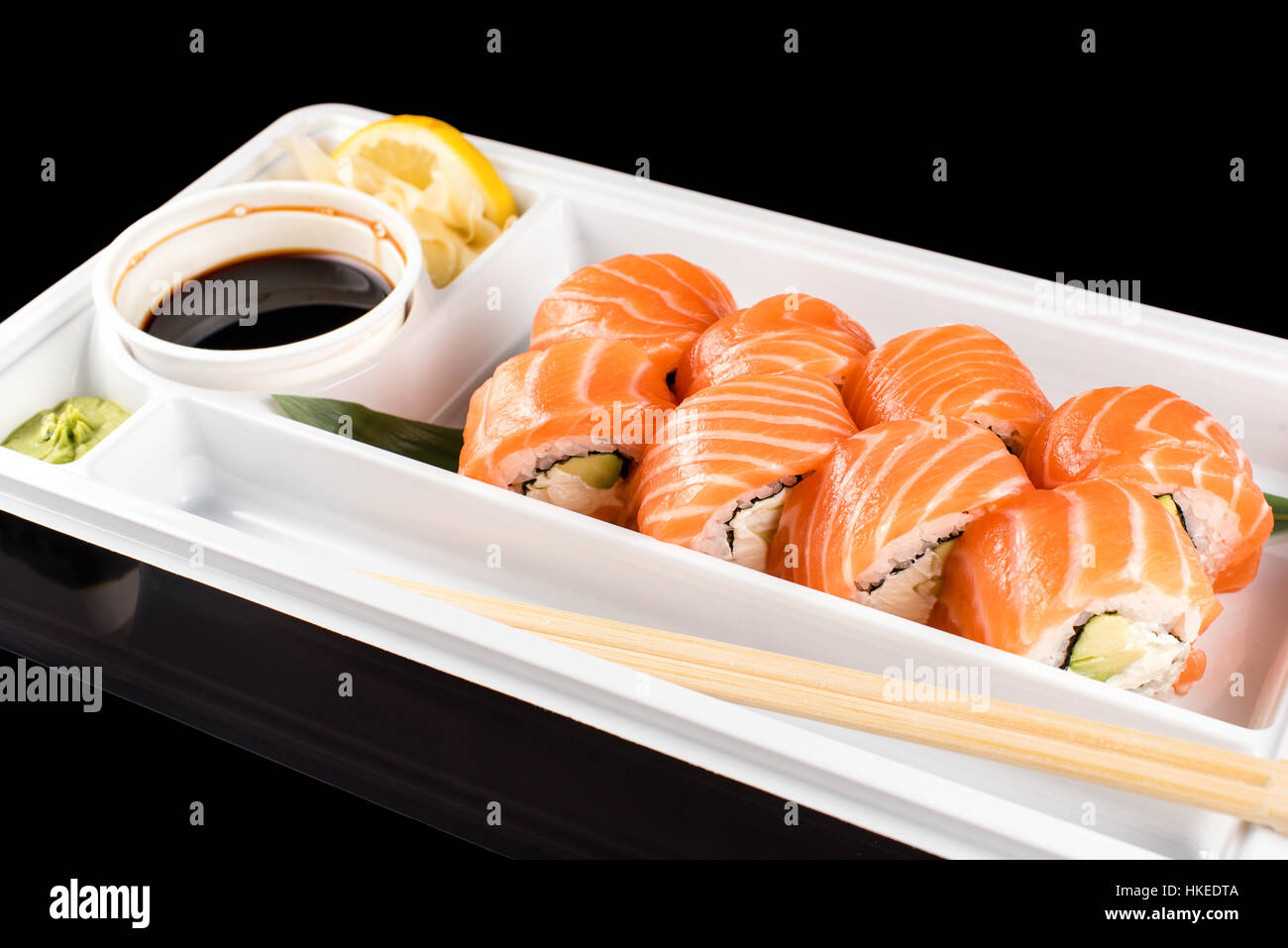 Sushi-Rollen von frischem rohen Lachs, Frischkäse und Avocado in weißem Kunststoff-Container bereit, auf schwarzem Hintergrund mit Reflexionen zu Essen gemacht. Stockfoto