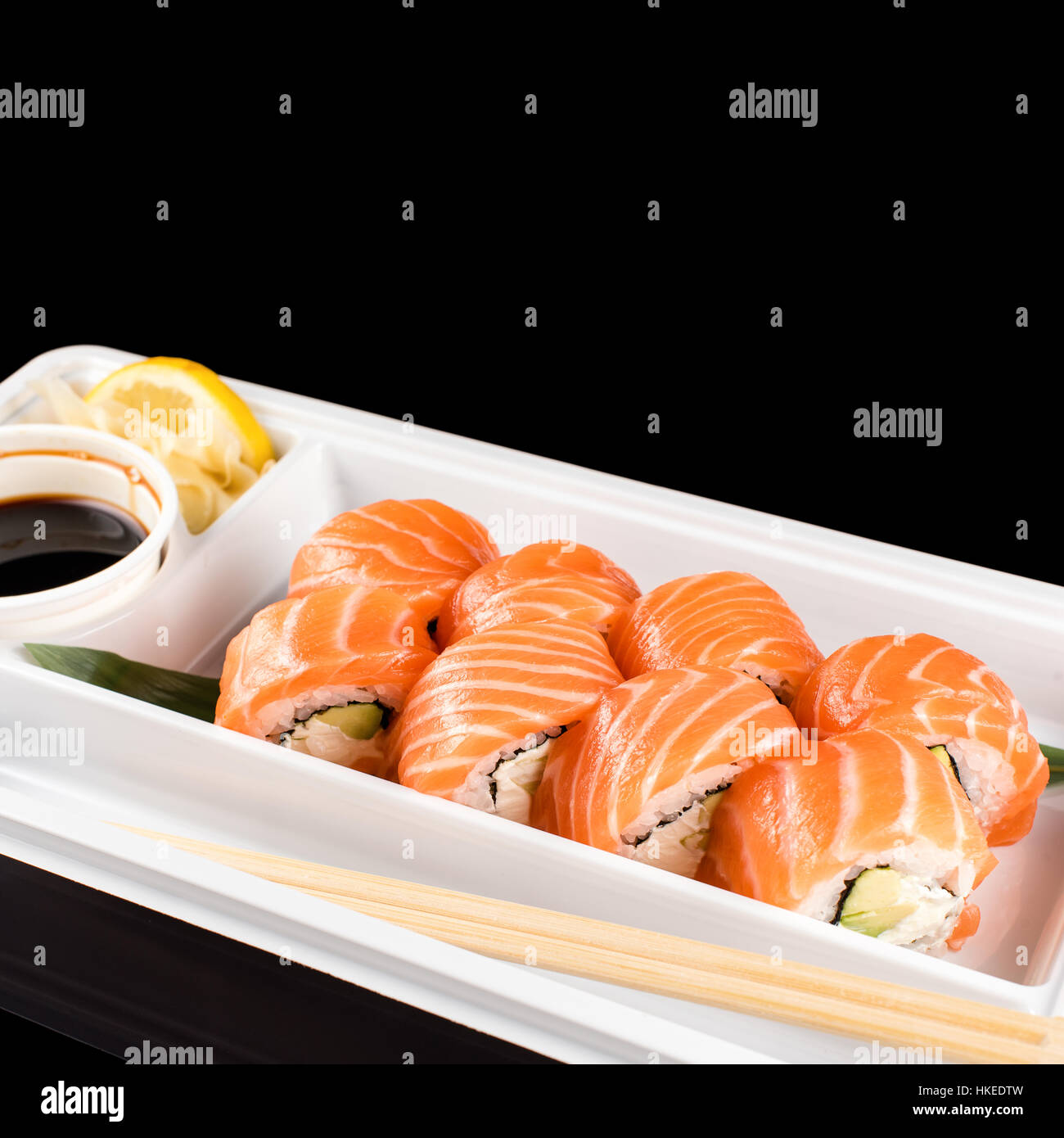 Sushi-Rollen von frischem rohen Lachs, Frischkäse und Avocado in weißem Kunststoff-Container bereit, auf schwarzem Hintergrund mit Reflexionen zu Essen gemacht. Stockfoto