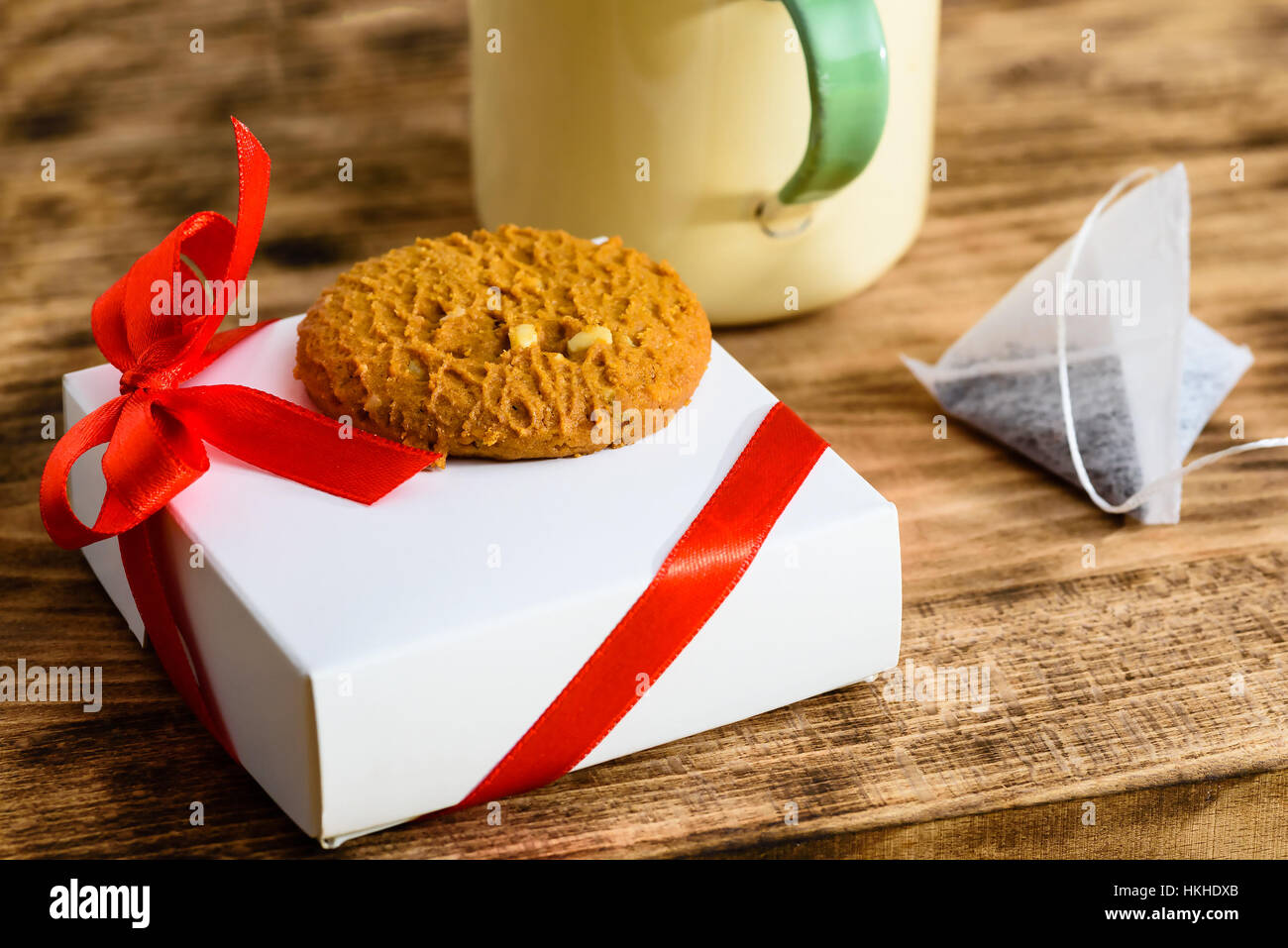 Kleine weiße mit roter Schleife und Cookie an der Spitze zusammen mit teilweise Teetasse und Teebeutel auf Tablett aus Holz. Cookie im Fokus Stockfoto