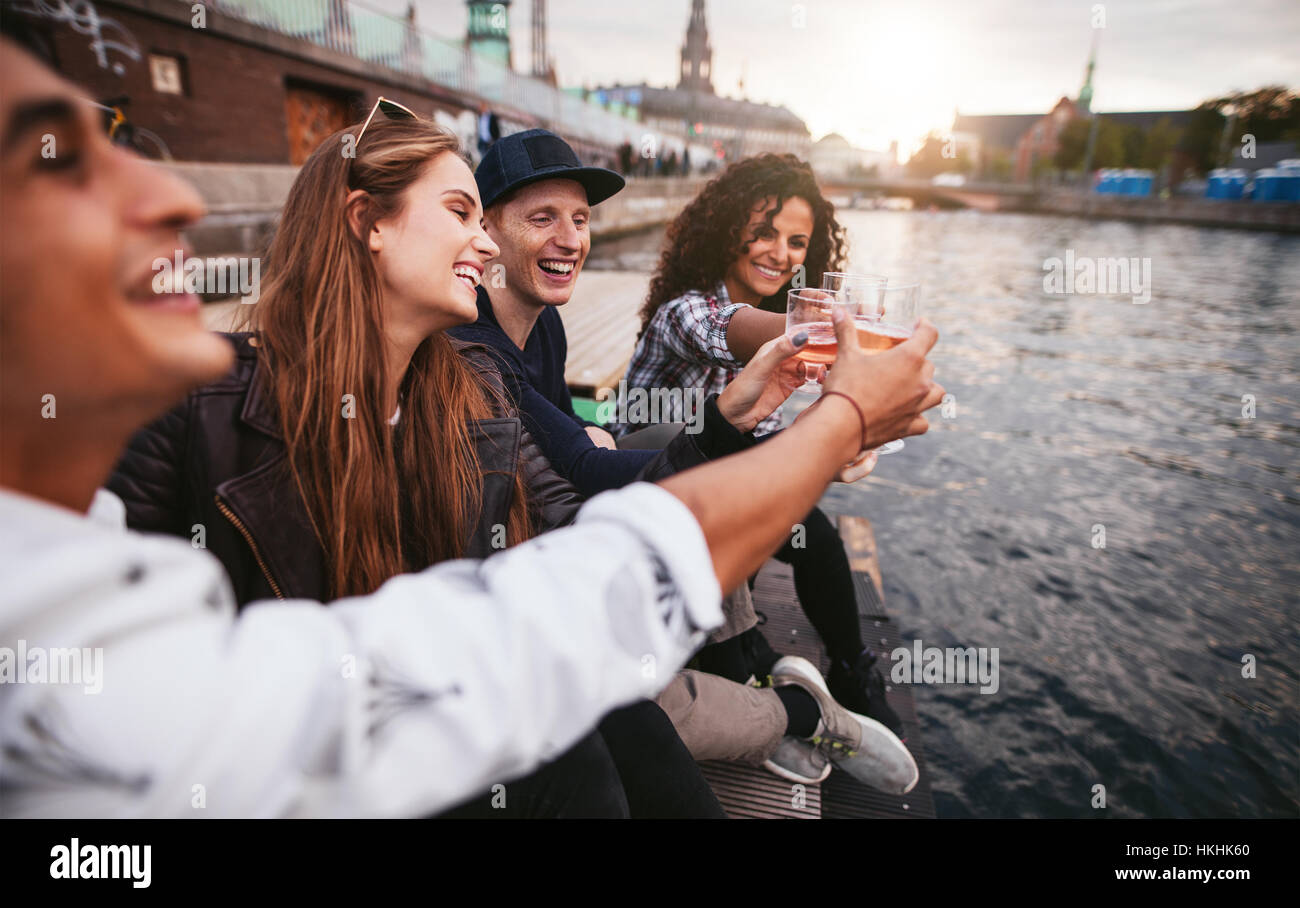 Gruppe von jungen Leuten Spaß im Sommer und mit Getränken zusammen. Sie sitzen auf einem Steg am See und Toasten Gläser. Stockfoto