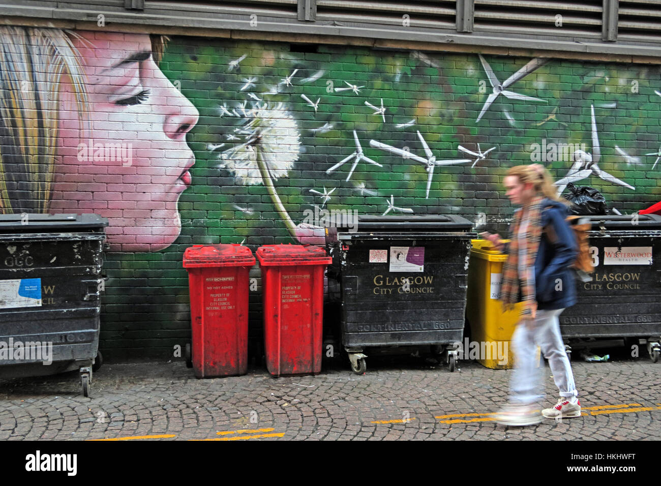 Graffiti in Glasgows Hinterstraße, blonde Frau bläst einen Löwenzahn in Windturbinen, erzeugt grüne Energie, in der Nähe von Mülltonnen Stockfoto