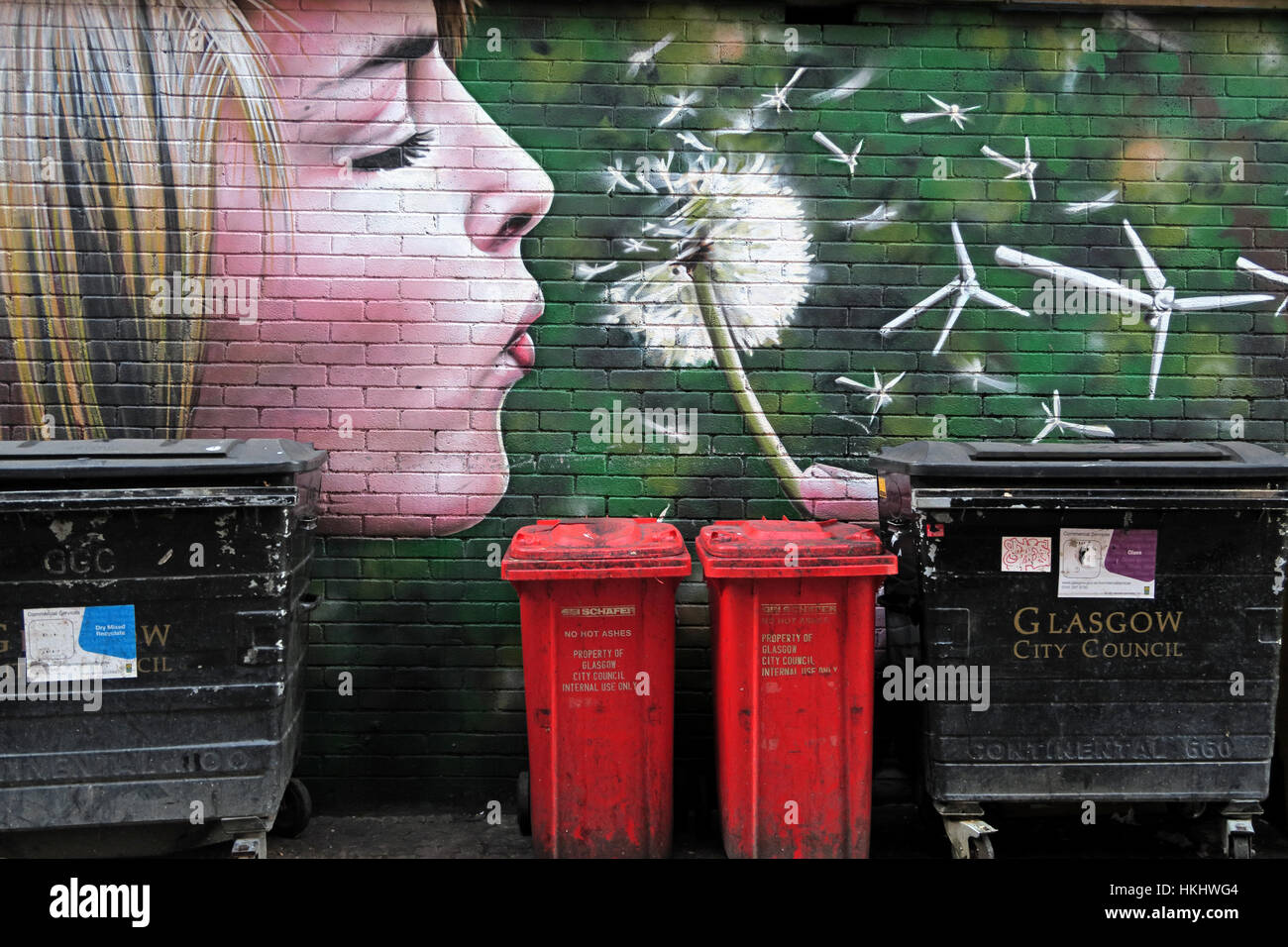 Graffiti in Glasgows Hinterstraße, blonde Frau bläst einen Löwenzahn in Windturbinen, erzeugt grüne Energie, in der Nähe von Mülltonnen Stockfoto