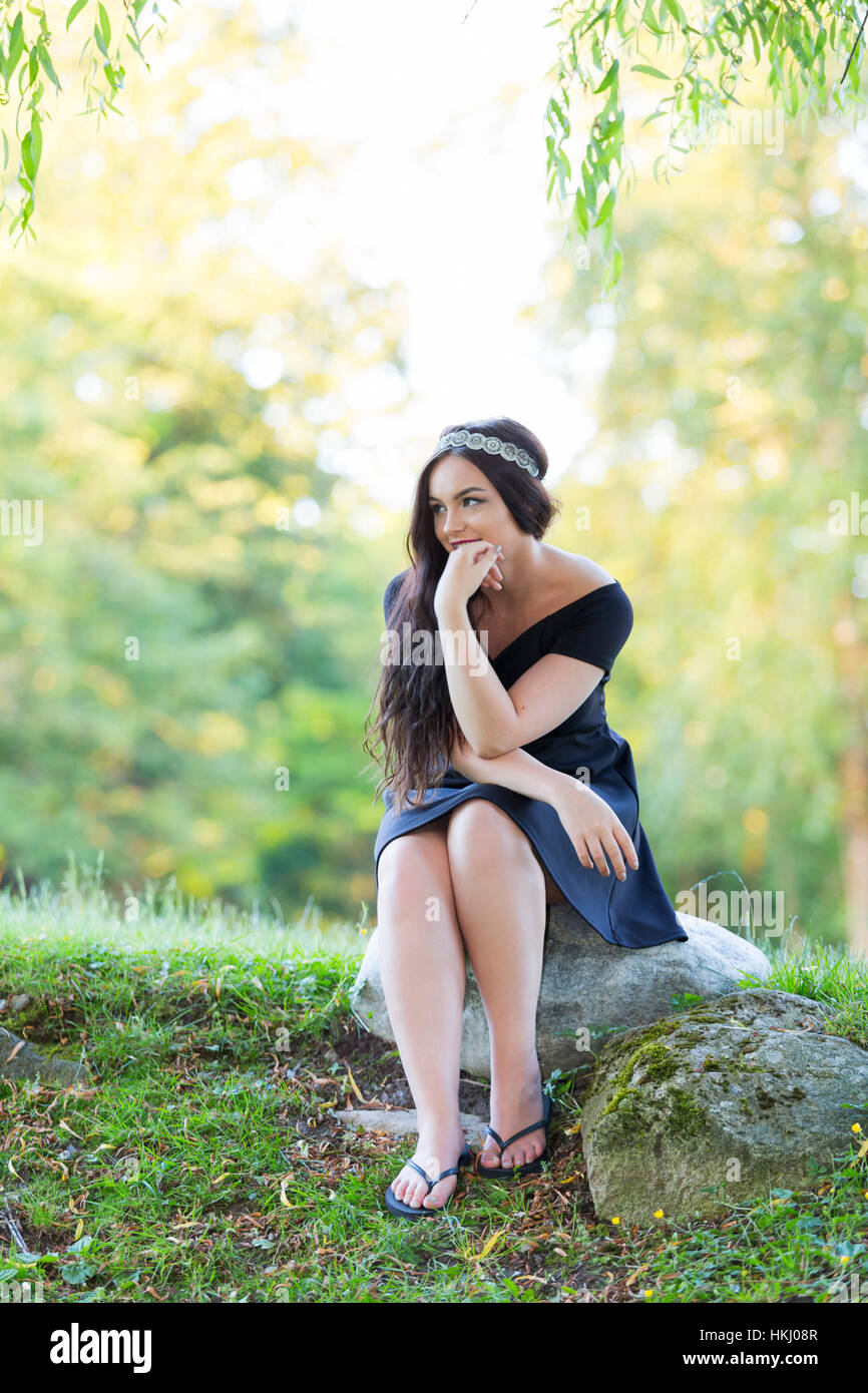 Ein junges Millennial Teenage Girl posiert für ihre Grad Porträts in Einem Park mit Einem funkelnden Stirnband und bescheidenen schwarzen Kleid, sitzend auf Einem Rock... Stockfoto