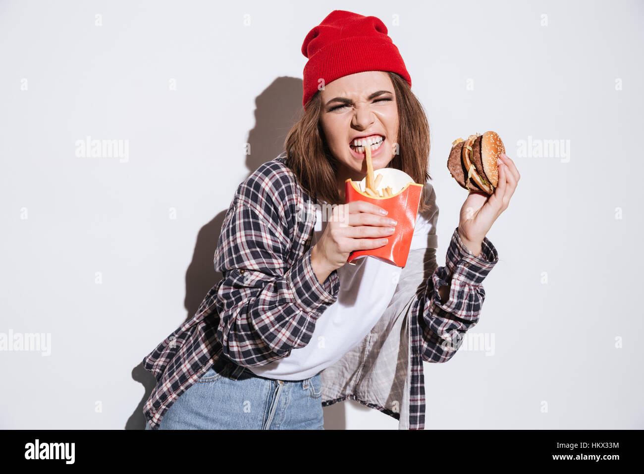 Bild von hungrigen böse Frau gekleidet im Hemd in einem Käfig Druck Hut überragt weißen Hintergrund isoliert und hält Pommes und burger Stockfoto