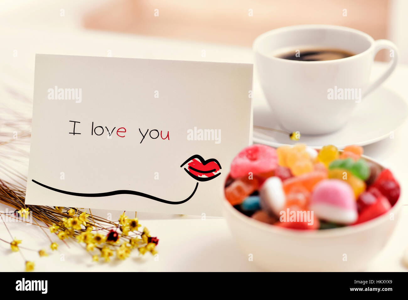 eine selbstgemachte Postkarte, gemacht von mir mit einem Kuss und den Text ich liebe dich, auf einem weißen Tisch neben einer Tasse Kaffee, Blumen und einem Bogen gezeichnet Stockfoto