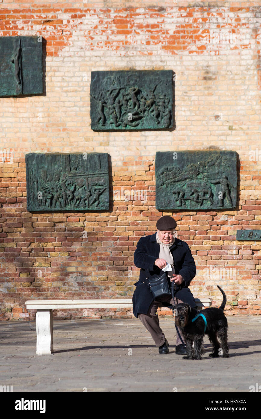 Mann, sitzend auf Bank mit Hund vor der Mauer der Erinnerung in der jüdischen Ghetto-Region von Cannaregio in Venedig, Italien im Januar Stockfoto