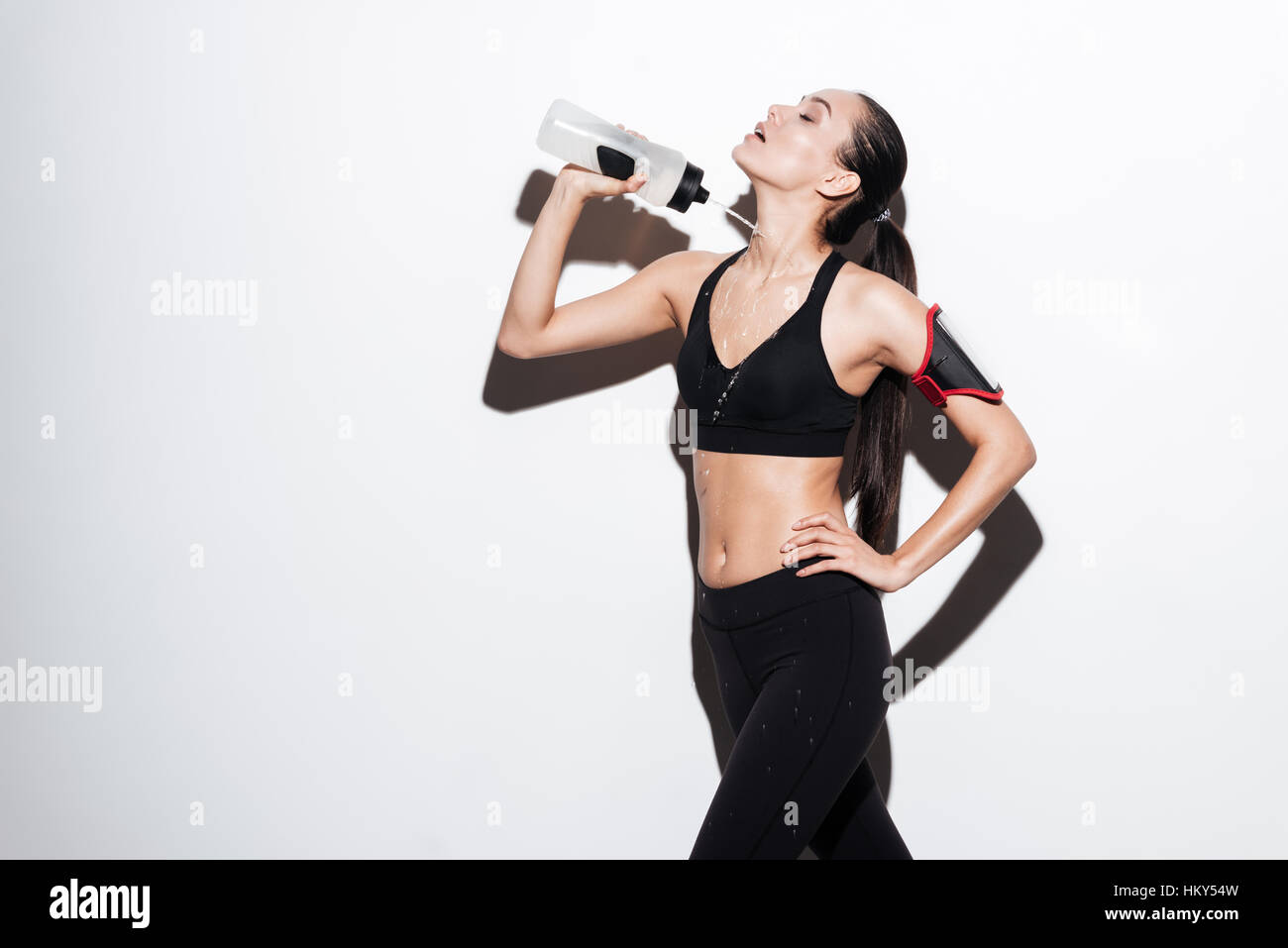 Verführerische junge Sportlerin gießt Wasser aus der Flasche auf sich selbst auf weißem Hintergrund Stockfoto