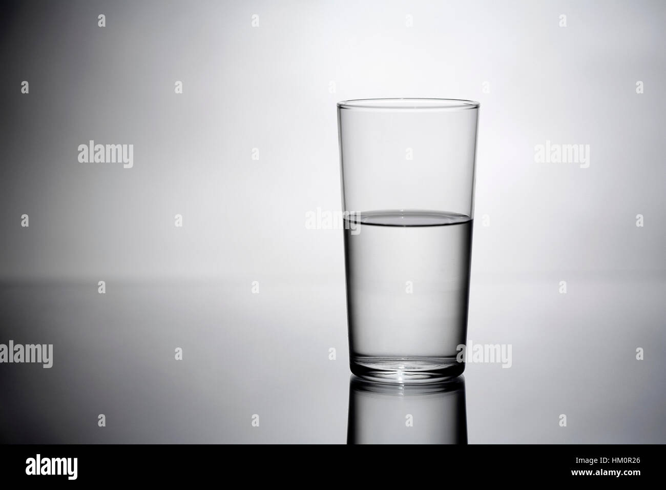 Ein halb volles Glas Wasser, das das Konzept illustriert, ist Ihr Glas halb voll oder halb leer, auf einer monochromatischen Reflexionsfläche Stockfoto