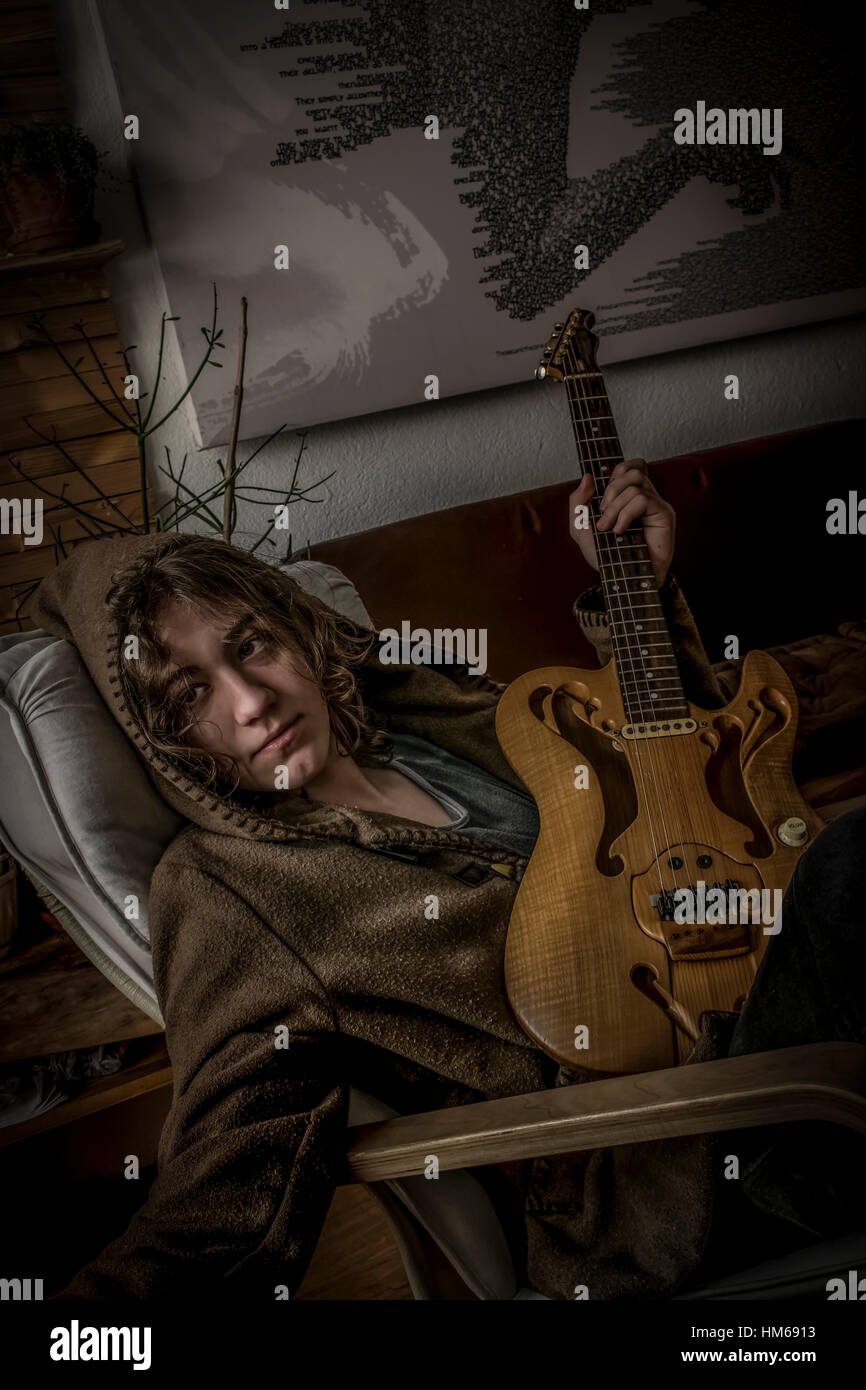 Ein junger Mann mit langen blonden Haaren und einer Kapuze, die auf einem Stuhl ruht und seine handgemachte halbhohle Gitarre in einem gemütlichen Ambiente hält. Stockfoto