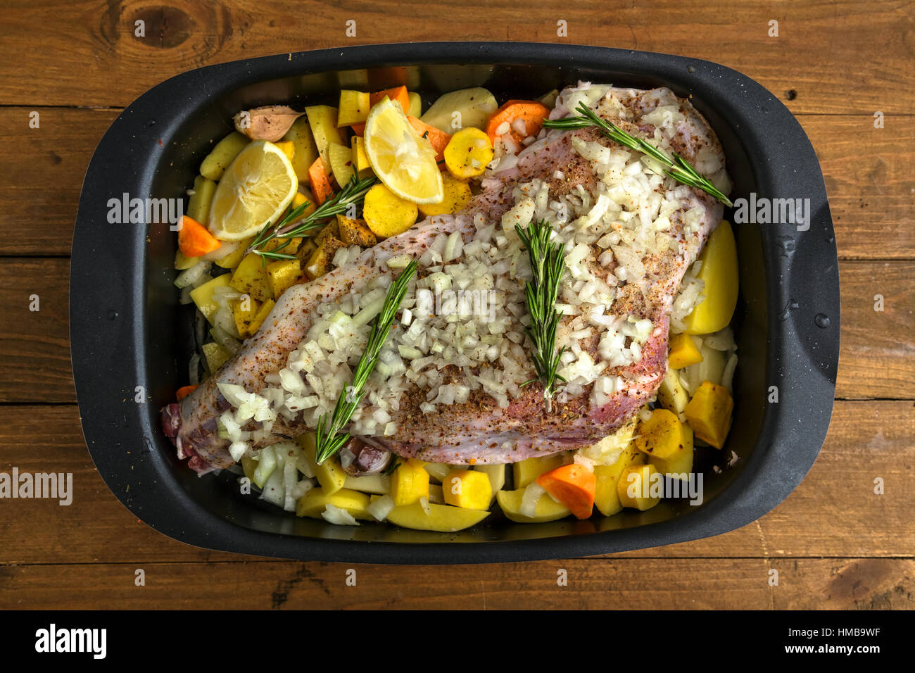 Rohe Lammkeule mit Wurzeln und Zwiebeln zum Braten auf einem rustikalen Holzbrett bereit. Ansicht von oben Stockfoto