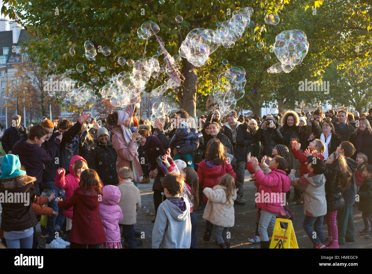 Kleine Kinder und deren Eltern versammeln sich um ein Straßenkünstler hinterließ eine Schneise der riesige Seifenblasen auf der Southbank in London Stockfoto