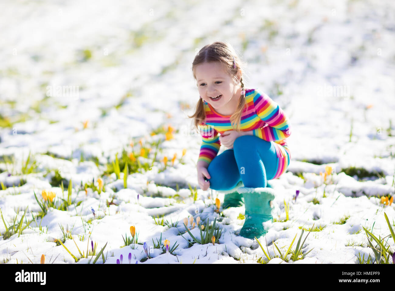 Niedliche kleine Mädchen in buntes Kleid, die ersten Blumen im Frühling Crocus unter Schnee an kalten Tag beobachten. Kind Kommissionierung Garten Blume Stockfoto