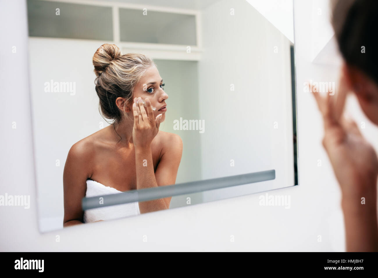 Reflexion einer Frau im Badezimmerspiegel kosmetische Creme im Gesicht auftragen. Frauen setzen auf Feuchtigkeitscreme auf ihre Gesichtshaut. Stockfoto