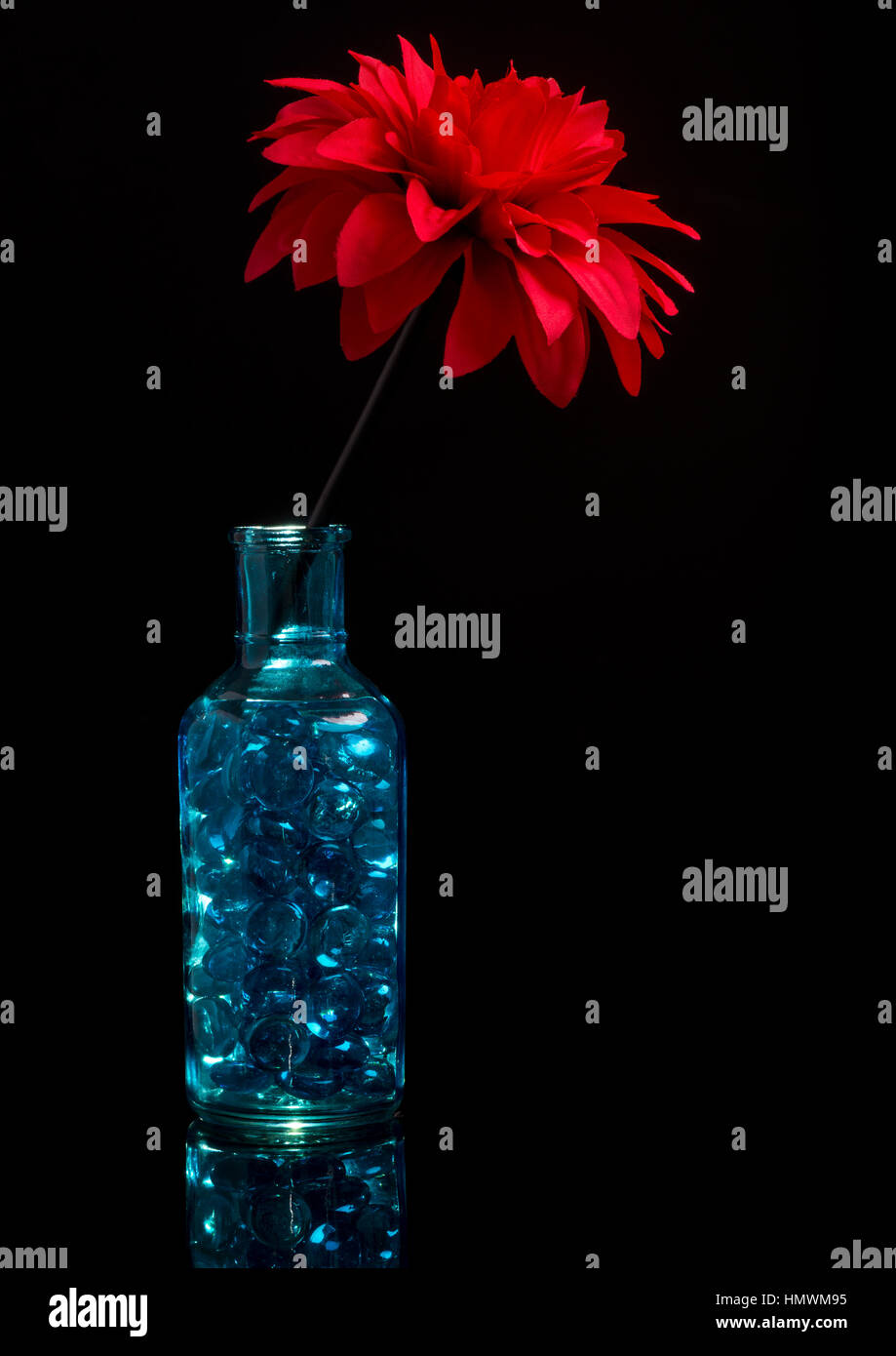 Eine blaue Glasflasche mit blauen Steinen, saß auf schwarzem Plexiglas. Eine künstliche rote Blume von oben.   Strobist: Die Blume war von meinem neuen Fa beleuchtet. Stockfoto