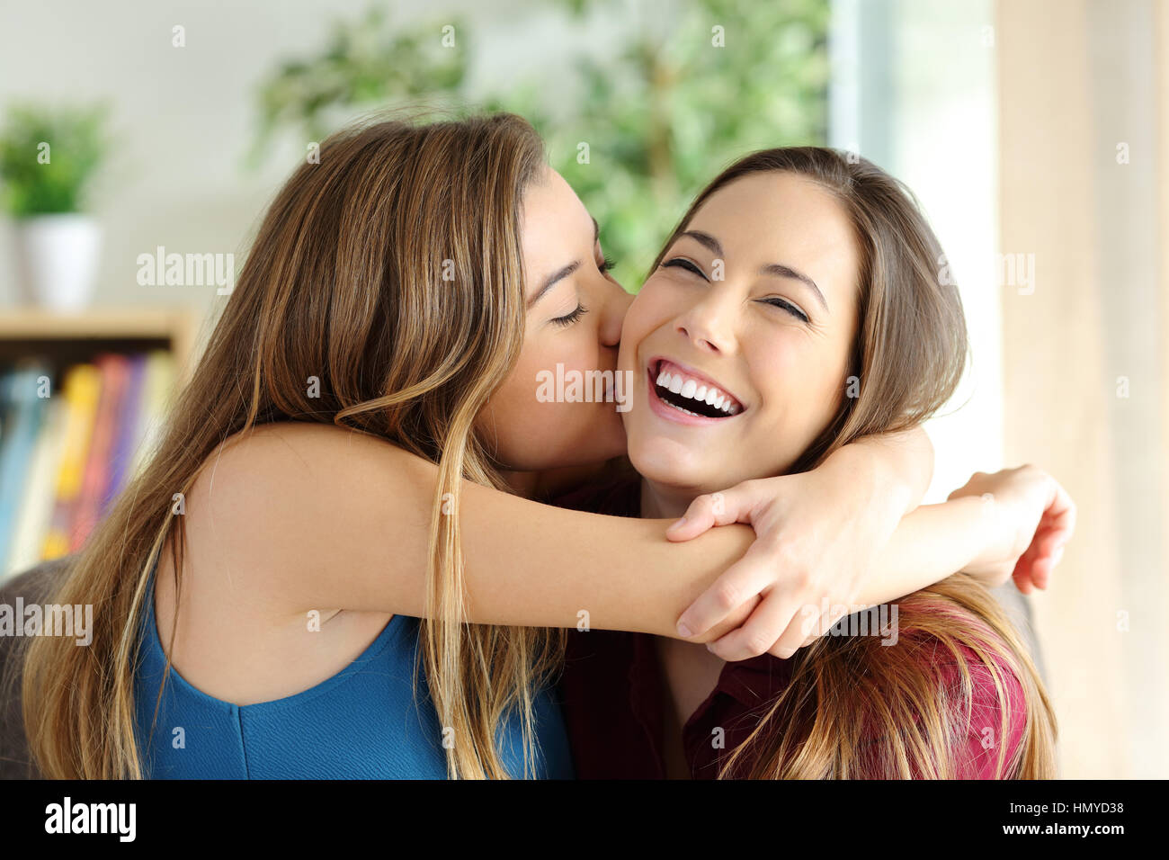 Liebevolle Mädchen küssen ihre glückliche Schwester oder Freundin im Wohnzimmer zu Hause mit einem heimeligen Hintergrund Stockfoto